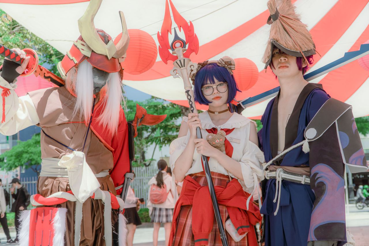 Three cosplayers dressed as fantasy characters pose together under vibrant canopy outdoors.