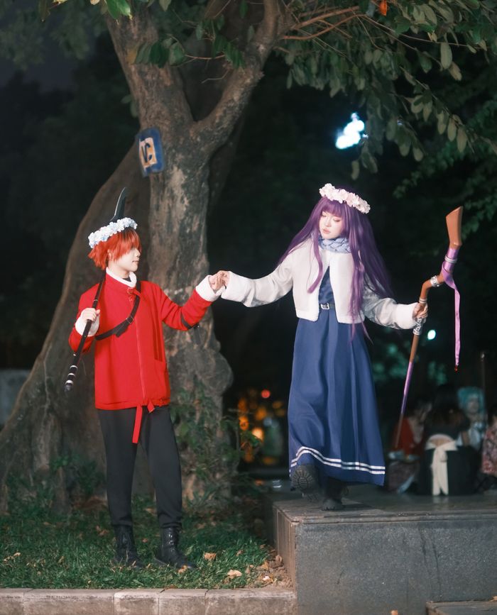 Two individuals in cosplay costumes pose under a tree at night, holding hands.